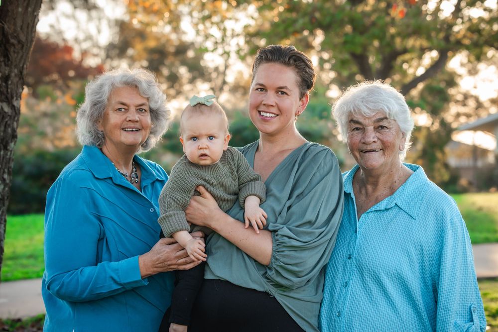 Four generations of women smiling together outdoors, with a young mother holding her baby between a grandmother and great-grandmother.