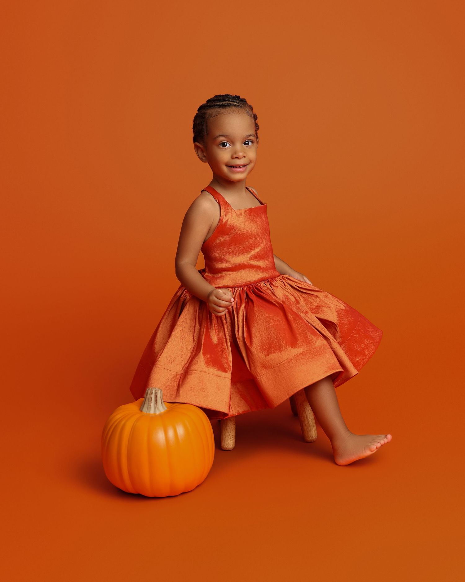 Young girl in orange dress sits on stool beside pumpkin against orange background.