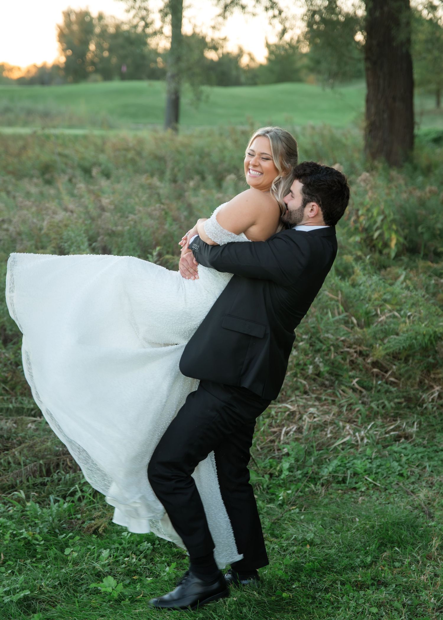 Groom in a black suit lifts and spins his smiling bride in a flowing white wedding gown on a grassy field at golden hour.