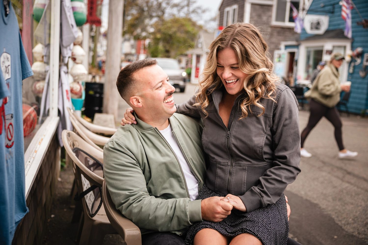 A couple laughs together while sitting on the steps of a food truck in a street festival setting.  Engagement at Rockport MA