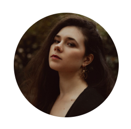 Circular portrait of a woman with long dark hair and gold earrings, wearing a black top and glancing over her shoulder against a dark green 