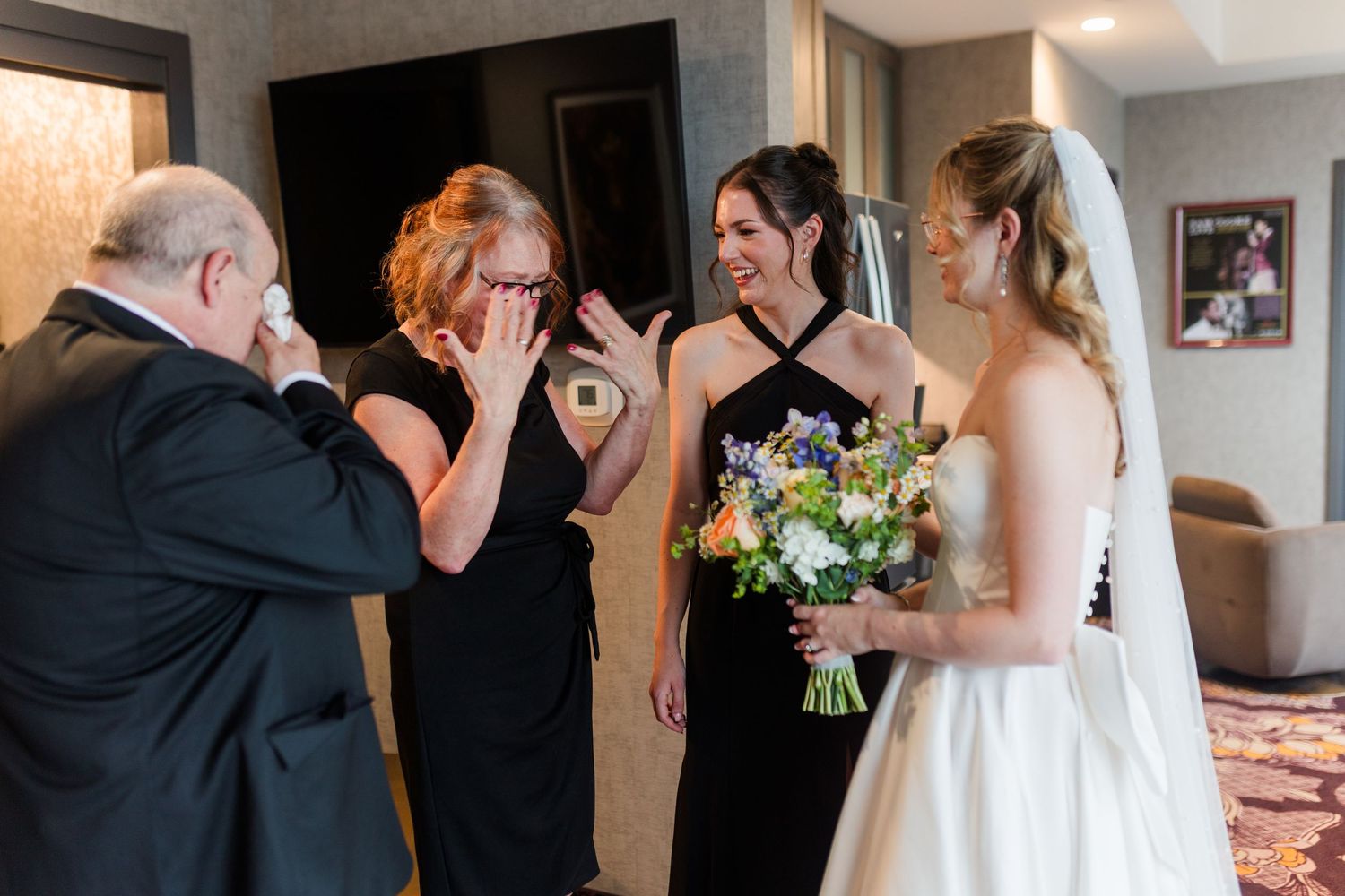 Emotional bride showing her wedding dress to tearful family members holding a colorful bouquet in a hotel room.