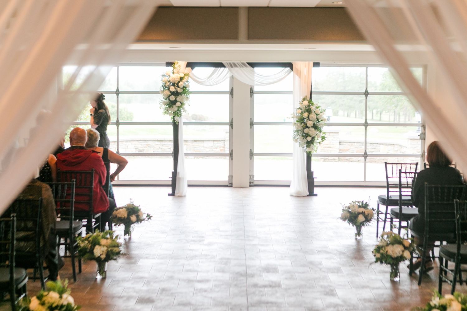 Elegant indoor wedding ceremony setup with floral arrangements, white drapes, and guests seated in dark chairs.
