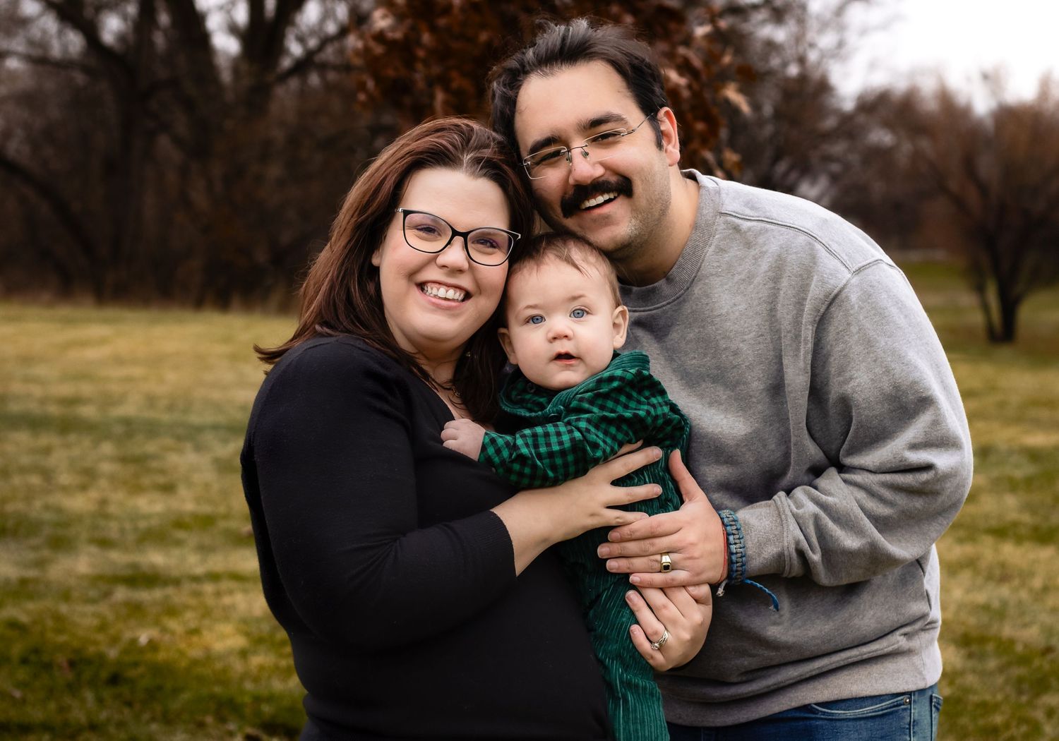 A family photo with the father and mother holding baby between them in a grass field.