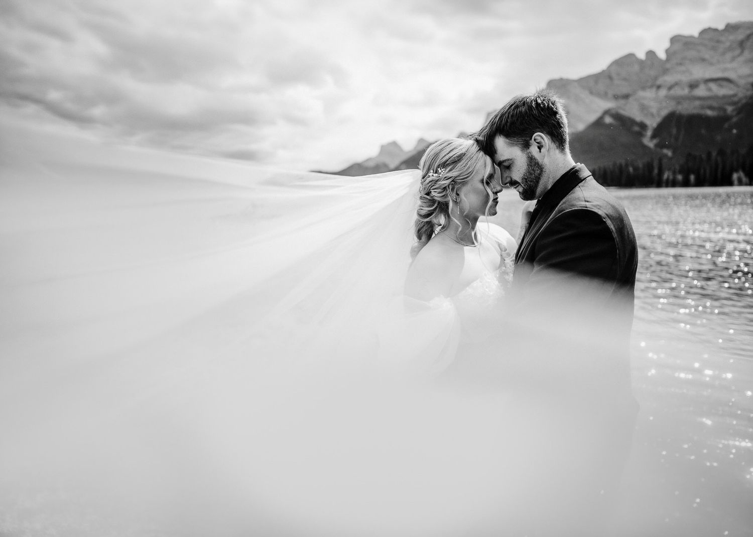 Black-and-white wedding portrait, couple forehead to forehead as the veil sweeps by the Rundle Forebay in Canmore