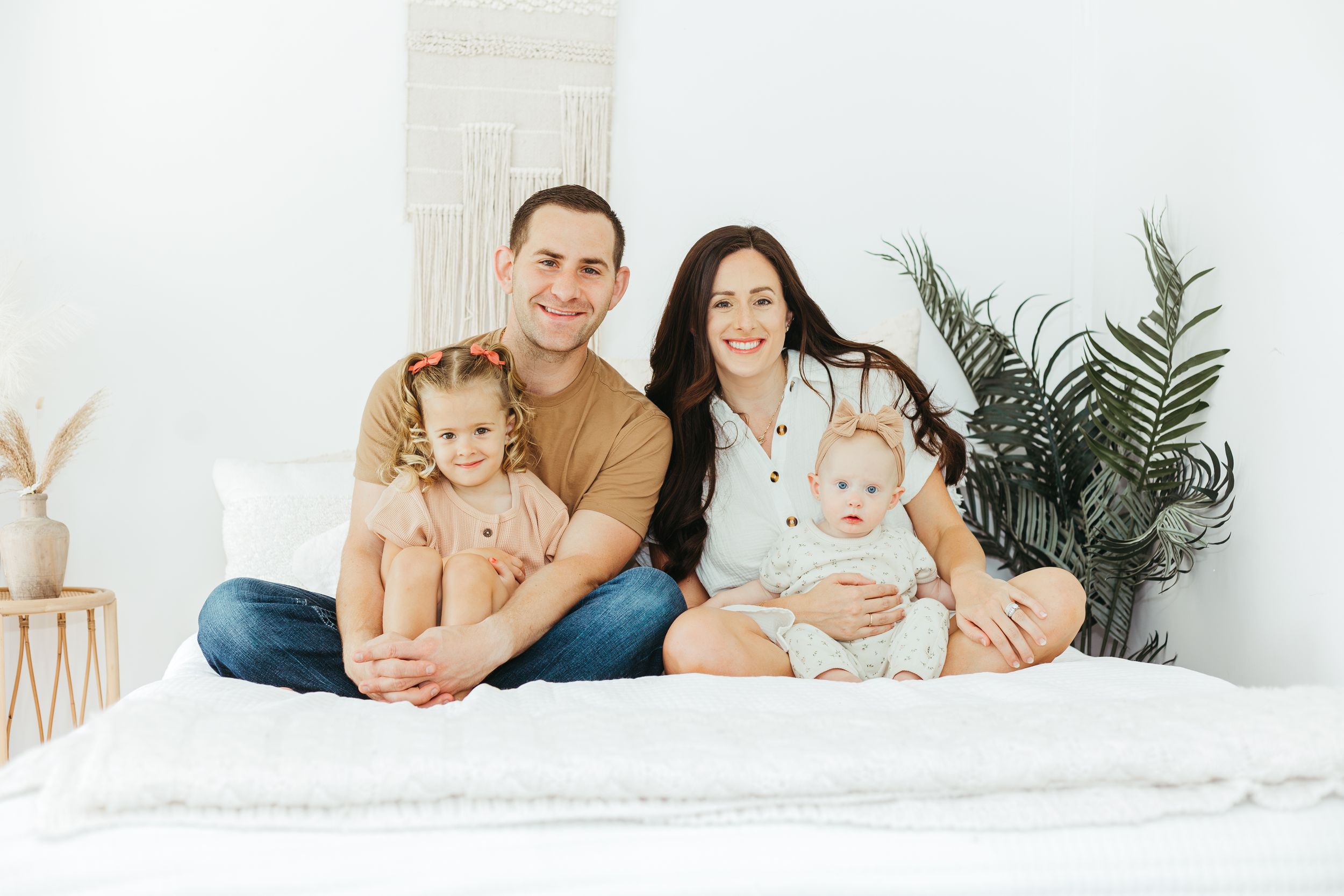 Happy family of four sitting on a white bed, smiling, with two young children in a bright modern room.