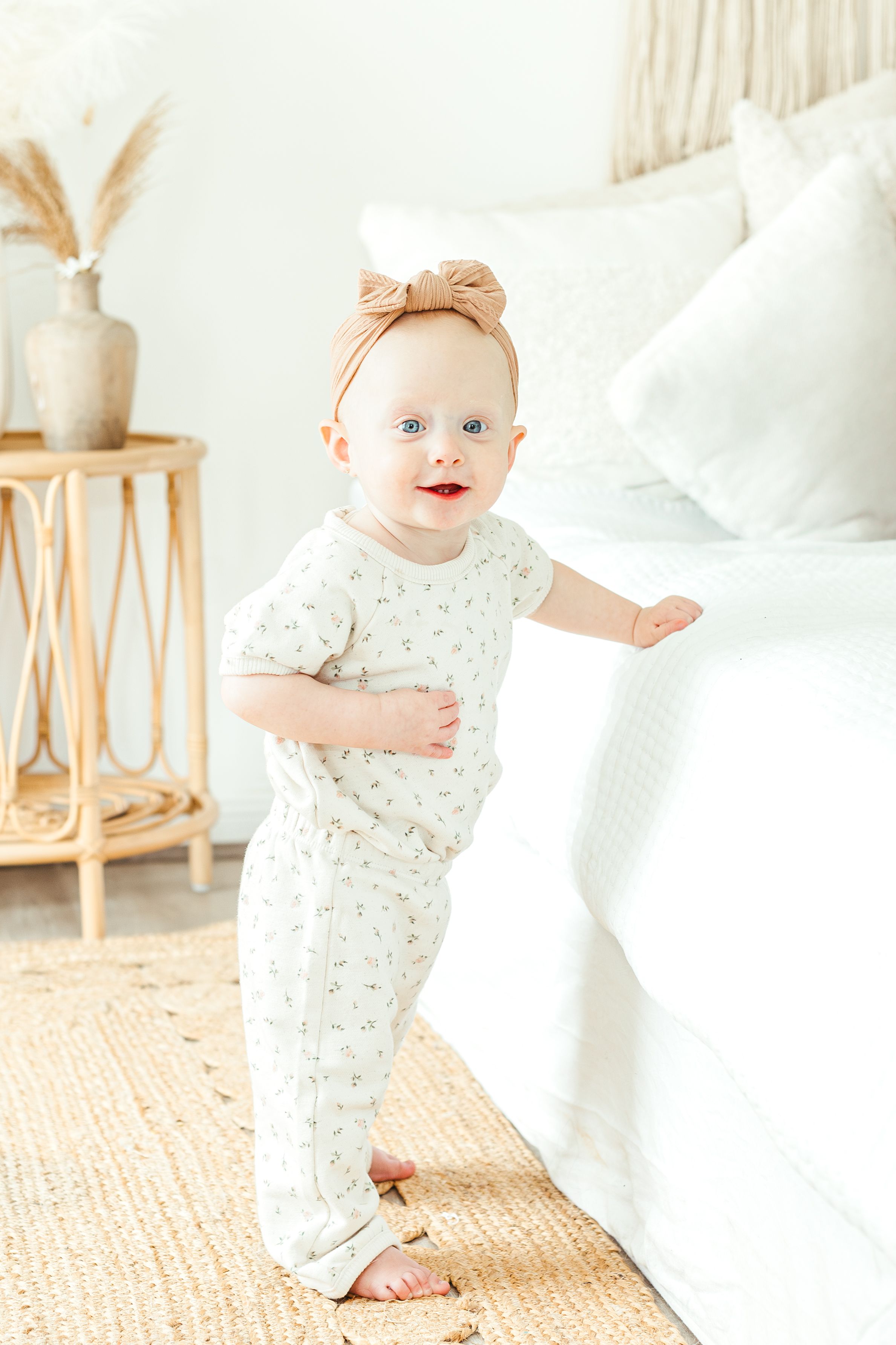 Adorable baby girl in floral romper and bow headband standing by a white bed in a bright nursery room.