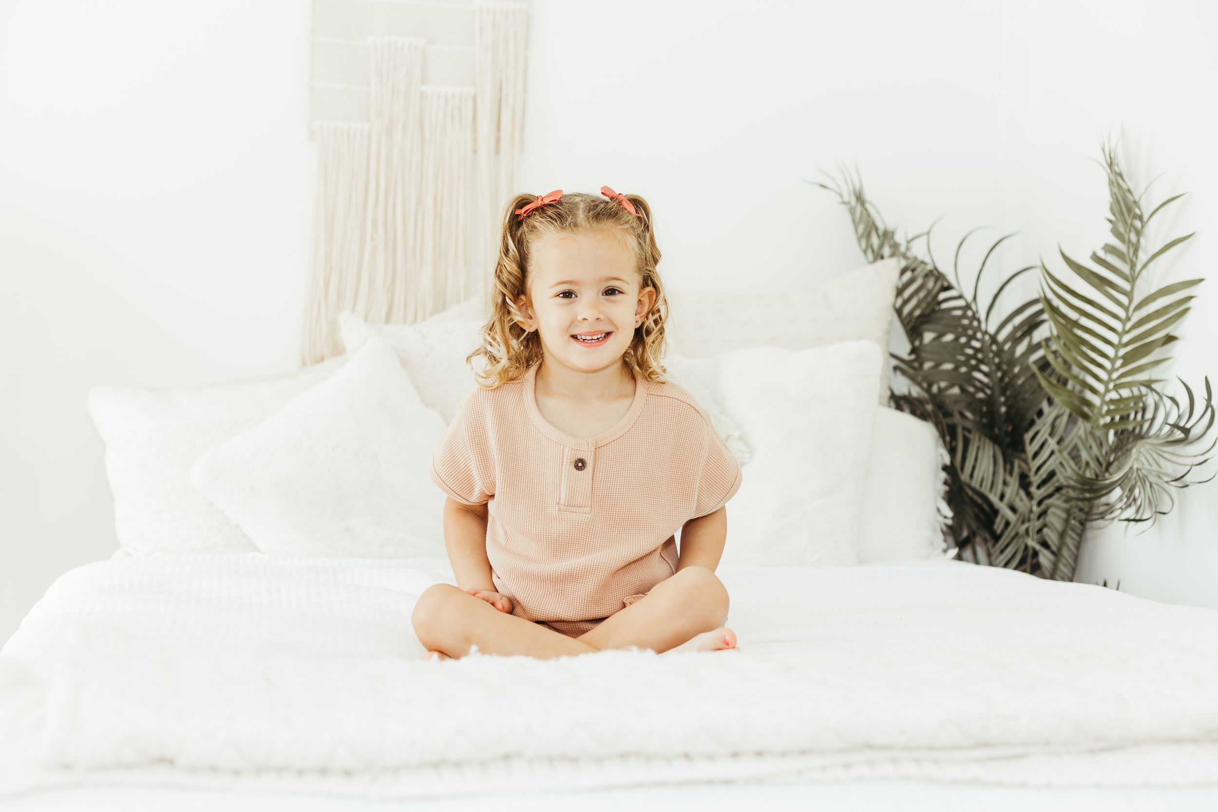 Young girl in pink outfit sitting cross-legged on white bed, smiling happily in bright white room.