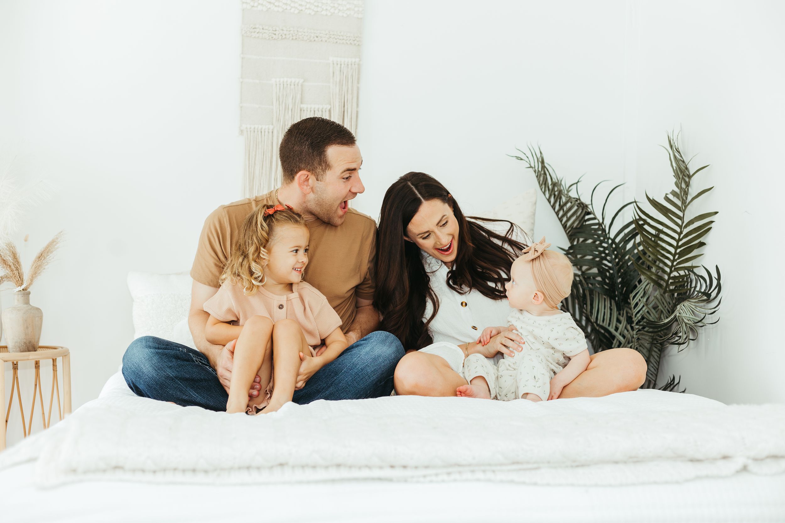 Happy family of four sitting on a white bed, parents smiling at baby while toddler sits nearby in bright room.