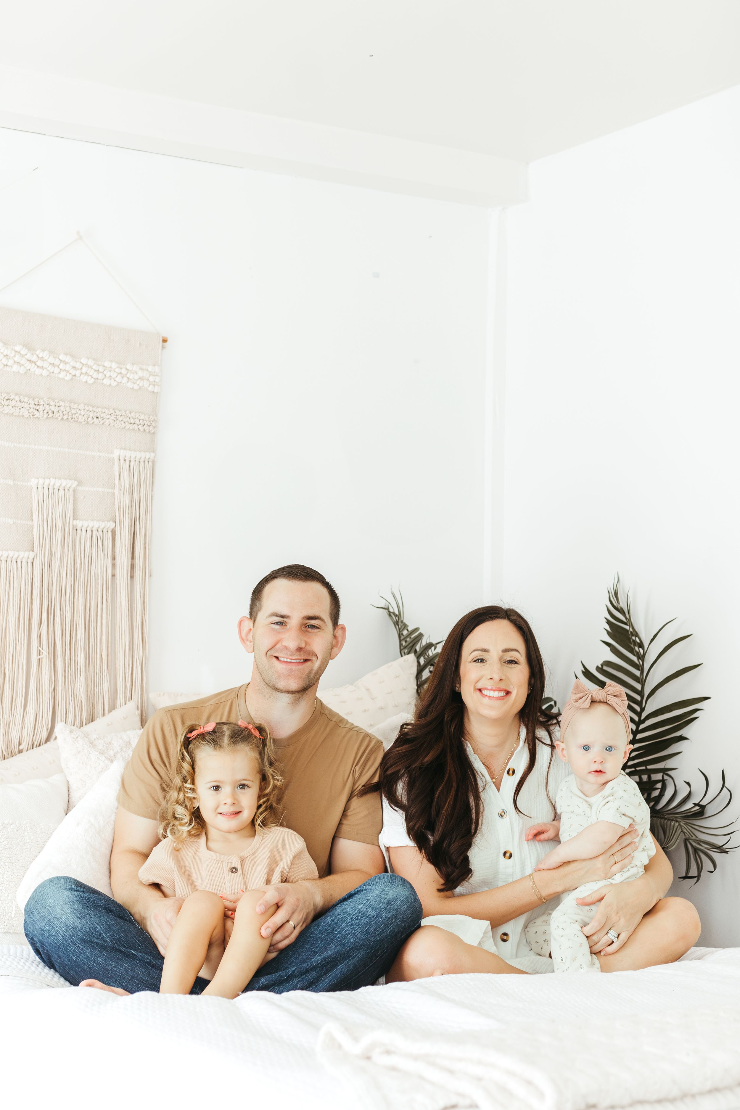 Happy family of four posing on a white bed in a bright boho-style room with macrame wall decor.