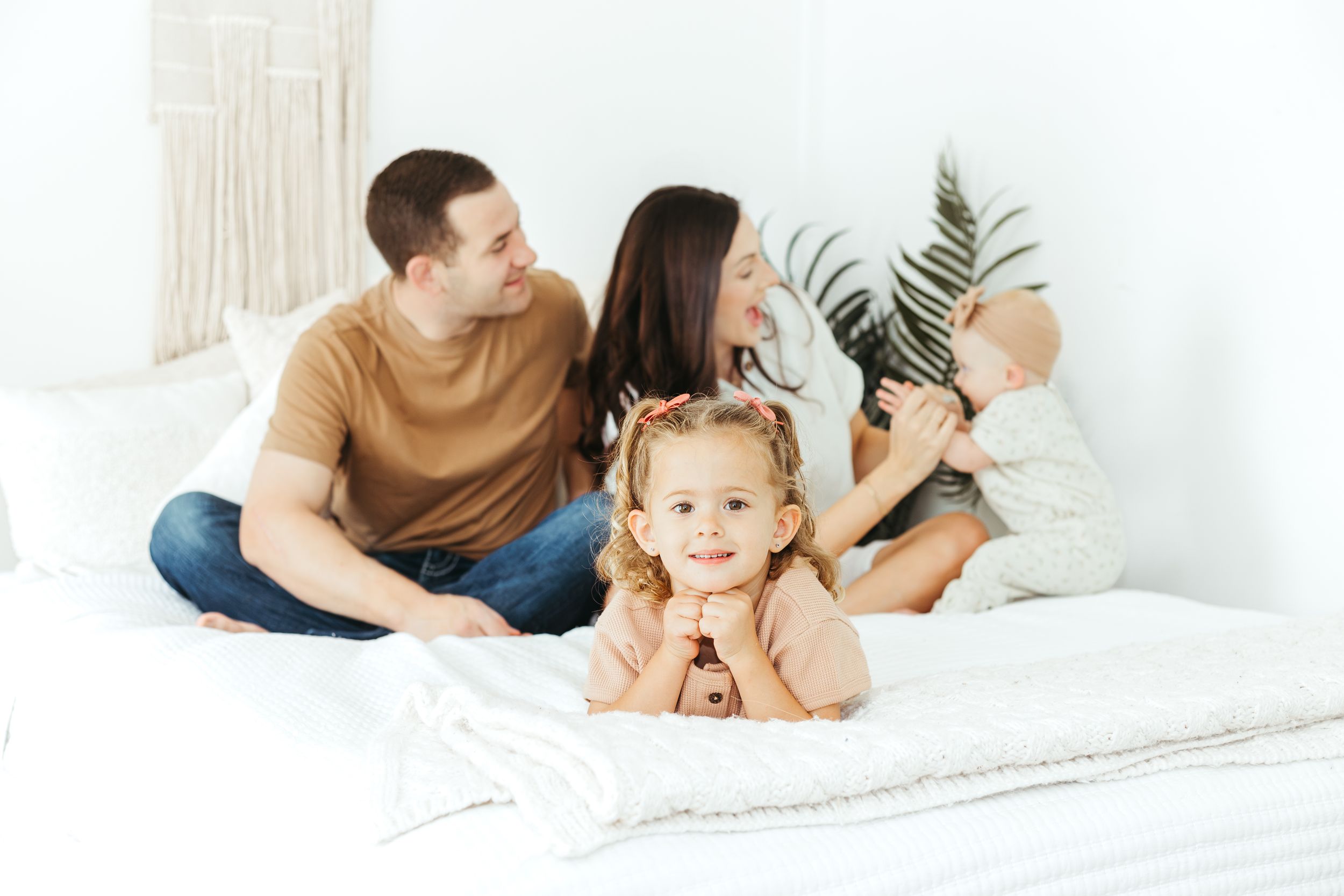 Happy young family with two children relaxing on a white bed in a bright, cozy bedroom setting.