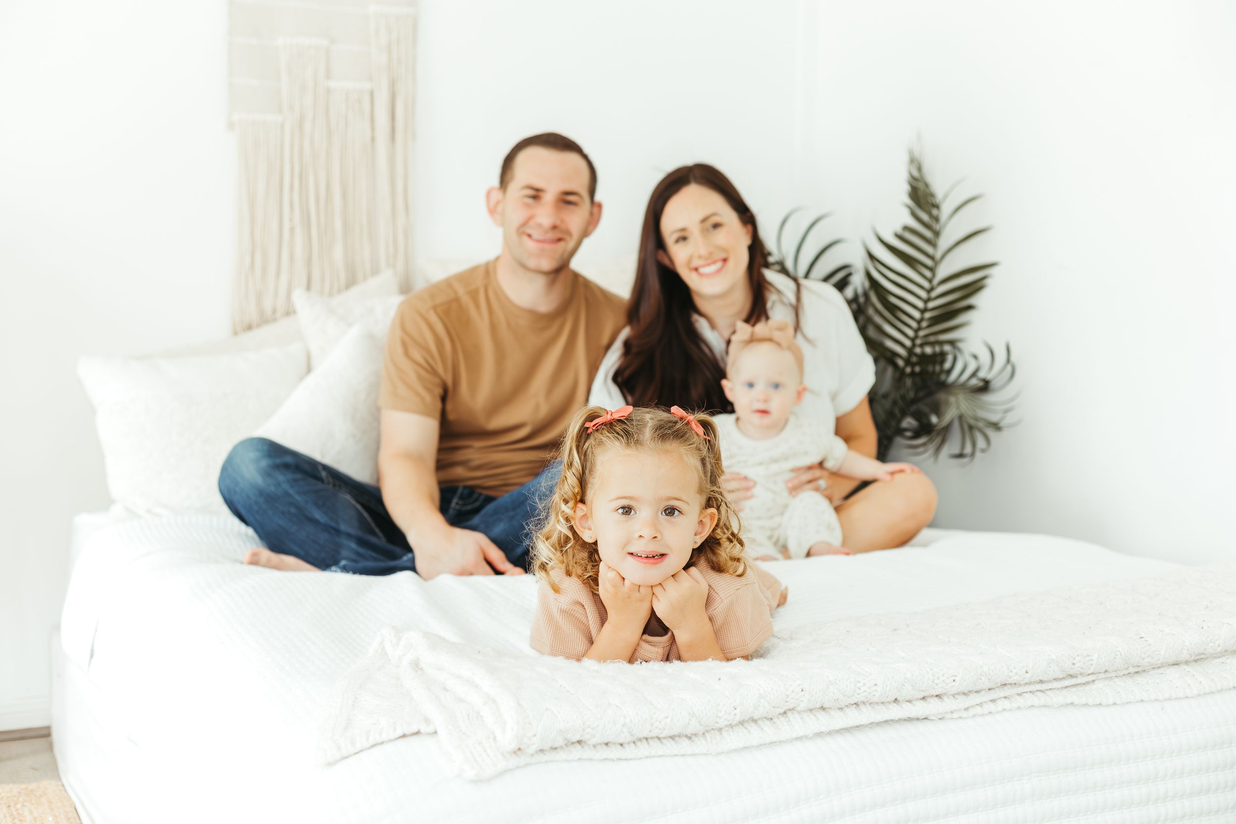 Happy family of four posing on a white bed, parents sitting with baby while young girl smiles in bright room.