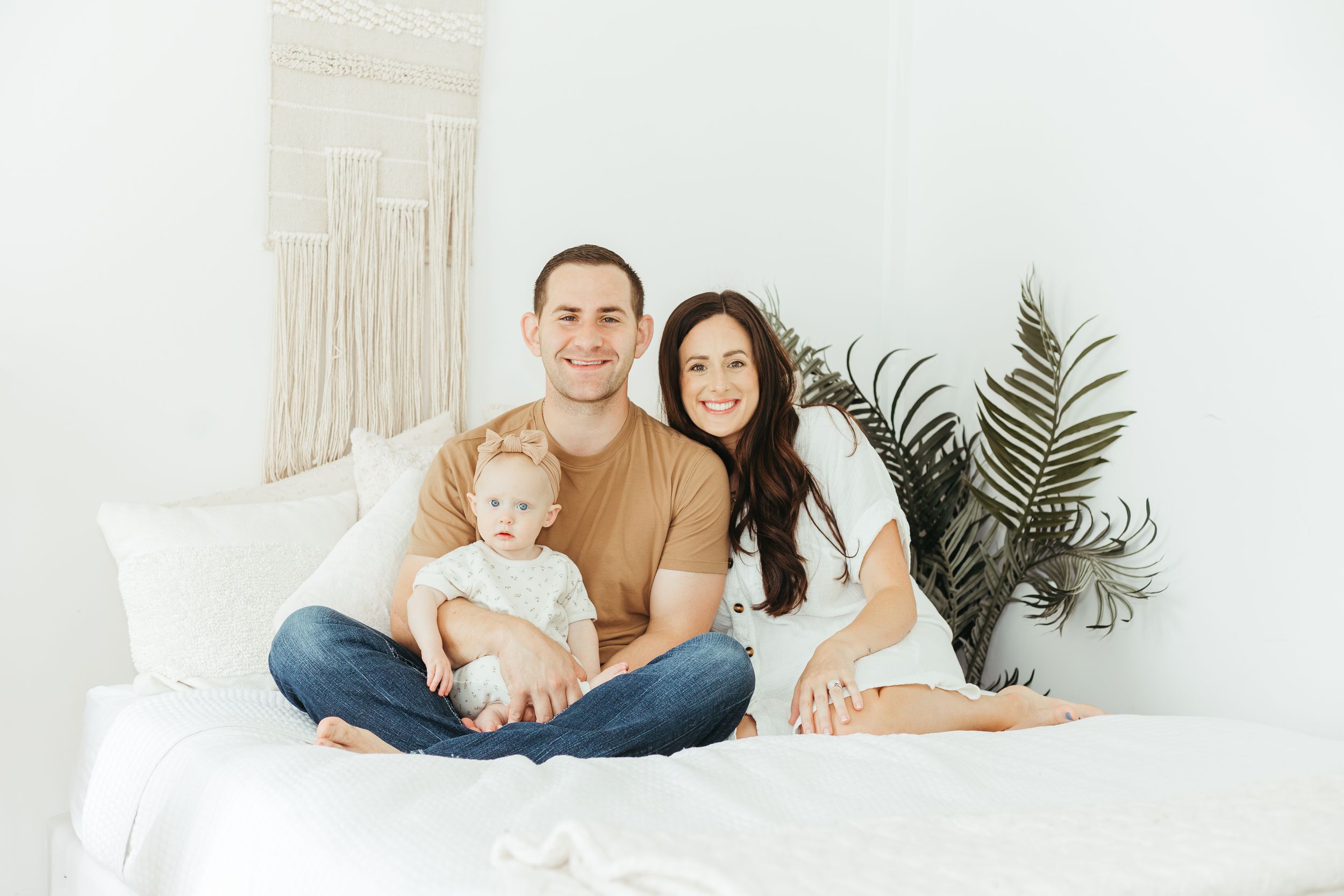 Happy young family with baby sitting on white bed in bright minimalist bedroom with plant decor.