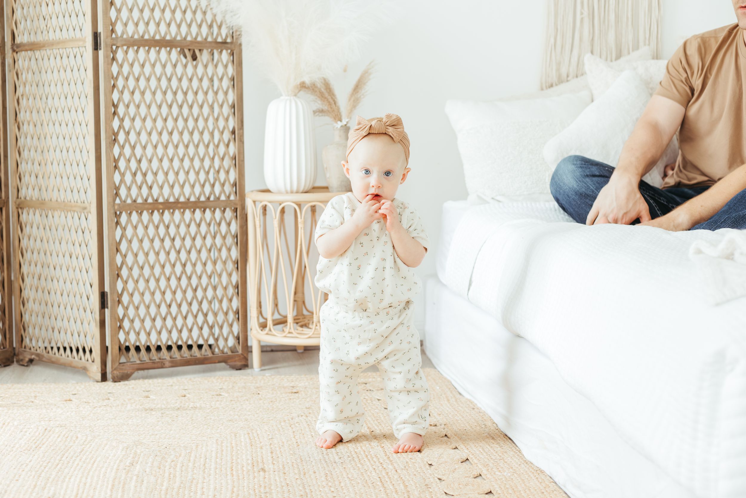 A toddler in a floral romper takes first steps in a bright boho living room while a parent sits nearby on a white sofa.