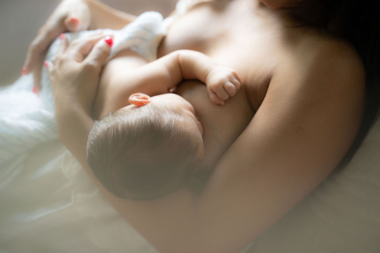 A mother tenderly holds her newborn baby against her chest in a soft, intimate breastfeeding moment.