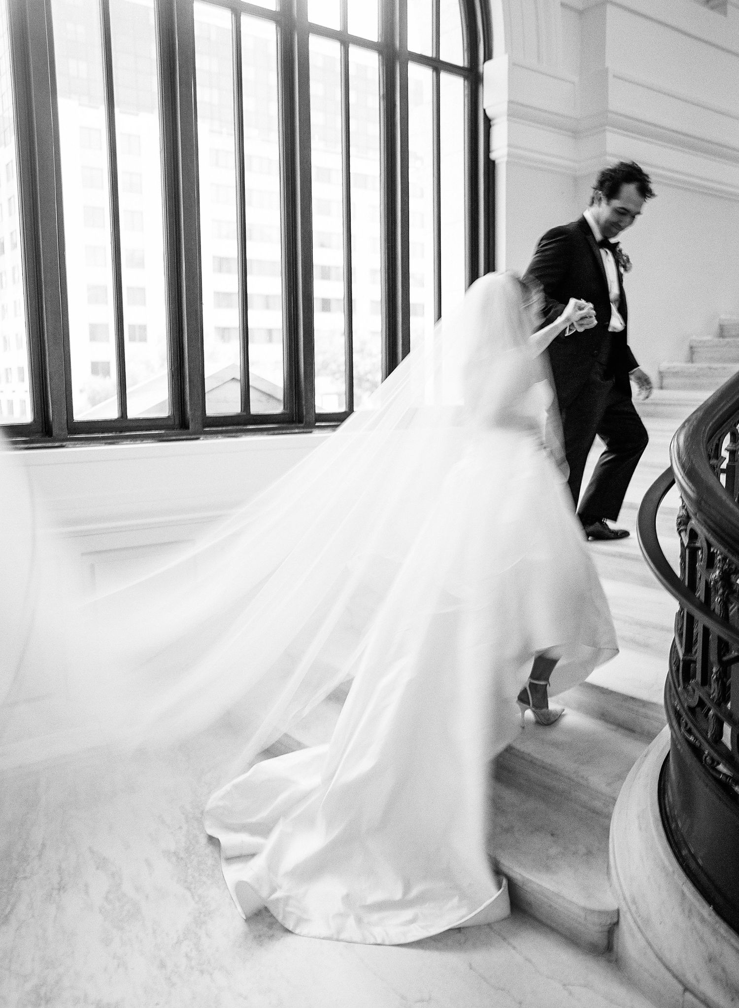 Black and white film photo of bride in flowing gown ascending marble staircase with groom in tuxedo in Washington DC