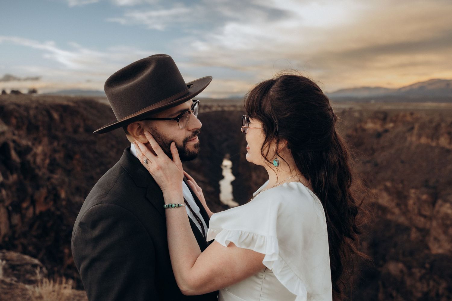 A romantic couple shares an intimate moment outdoors, man in black hat and blazer, woman in white dress, scenic canyon backdrop.