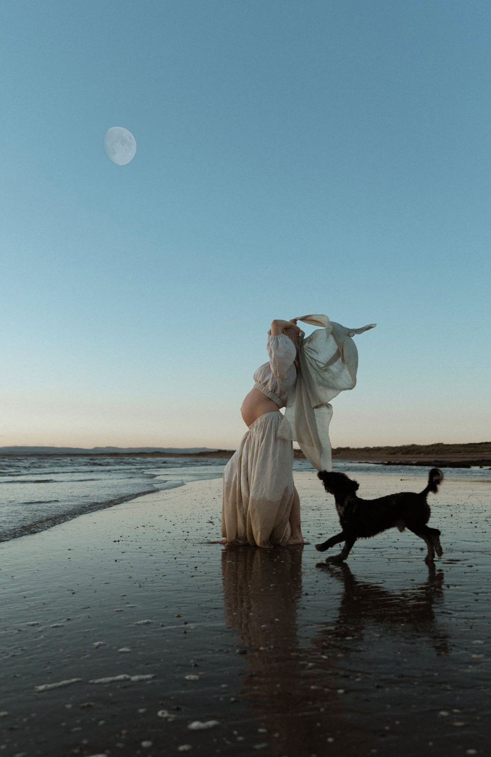 Pregnant woman standing barefoot on the shoreline at dusk, walking with her dog along the water’s edge.