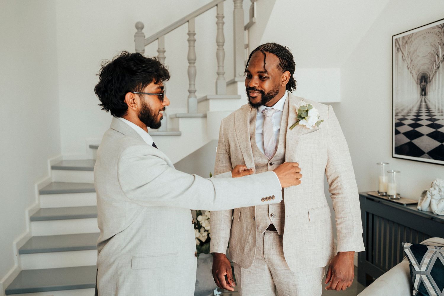 Two people in white shirts and suspenders getting ready for a formal event near a window.