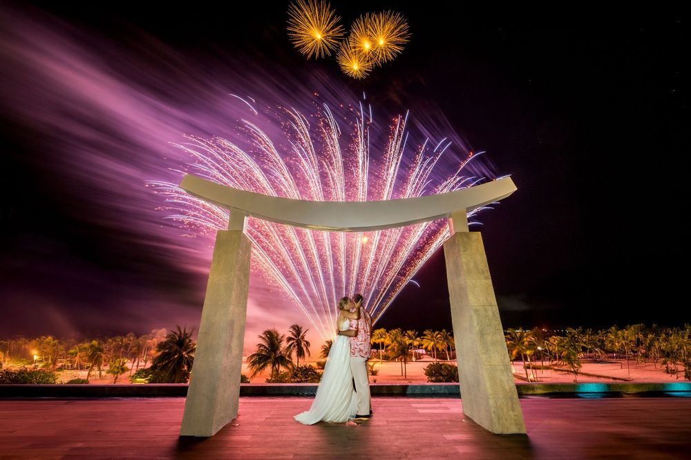 Fireworks illuminate the night sky over a beachfront wedding ceremony with a white draped arch and palm trees.
