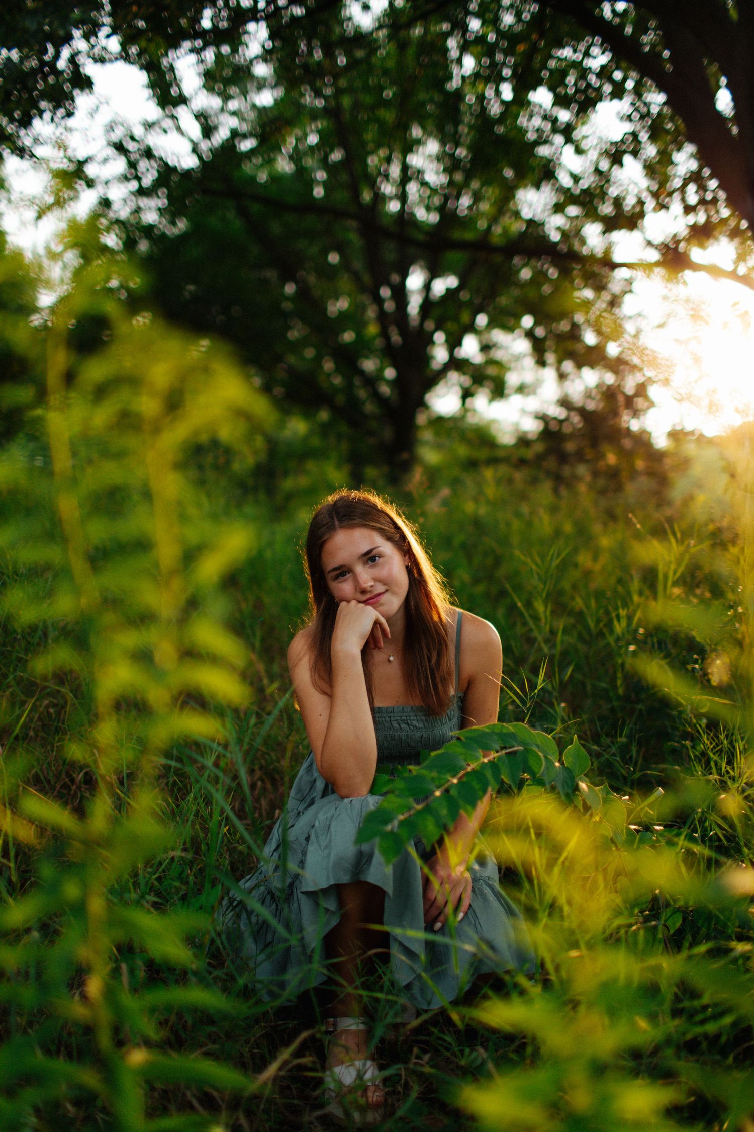 Person in dress poses thoughtfully among lush green foliage at sunset.