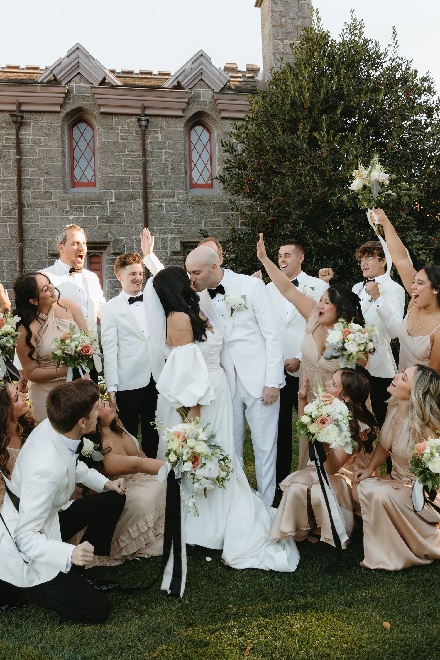 Wedding party in white attire poses playfully for group photos outside a stone building.