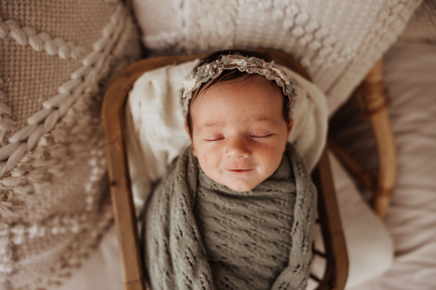 Sleeping newborn baby swaddled in sage green knit wrap wearing floral headband in wooden bowl.