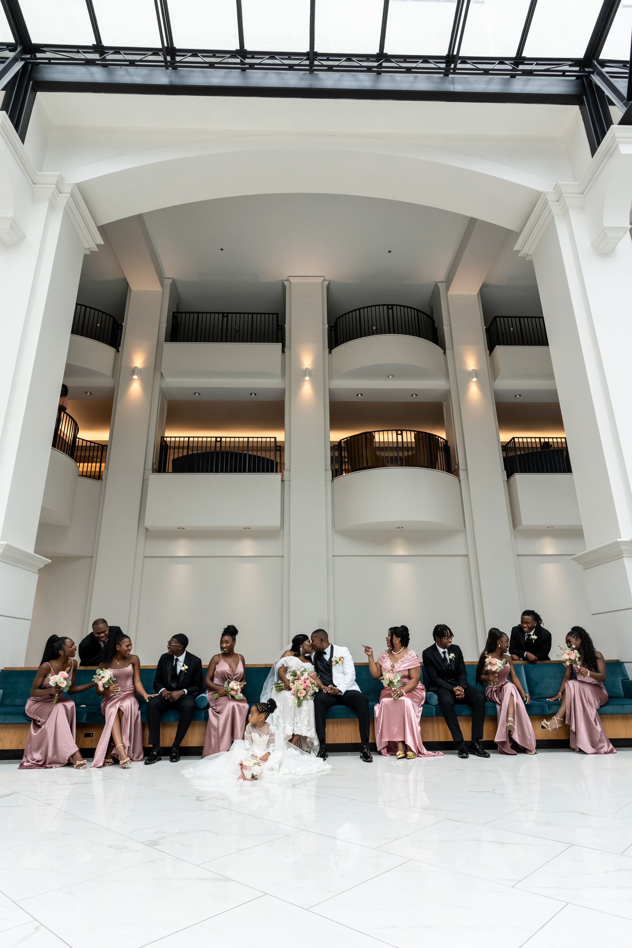 Wedding party members in pink and black attire pose creatively on white floor in elegant columned venue.
