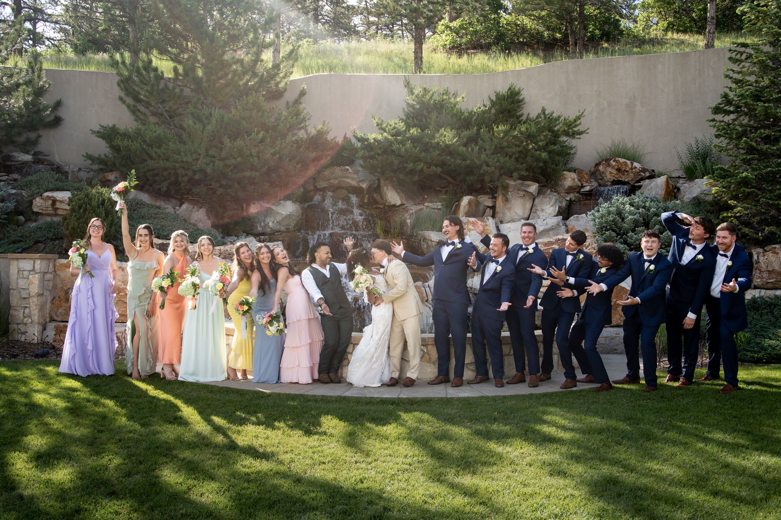 A large wedding party poses outdoors in a curved line formation under shade trees at a garden venue.