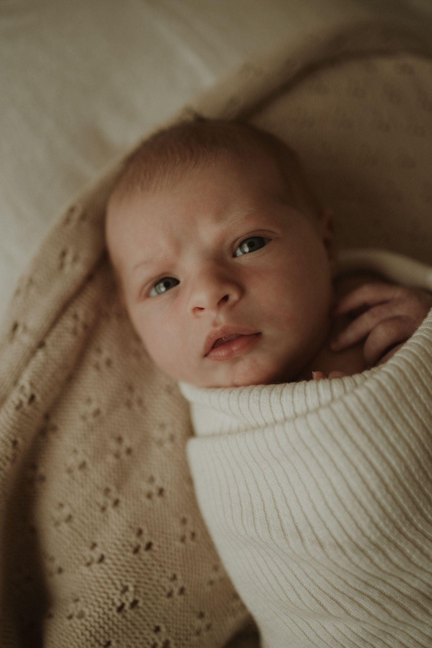 A small baby swaddled in white fabric rests on soft beige fabric with delicate eyelet details.