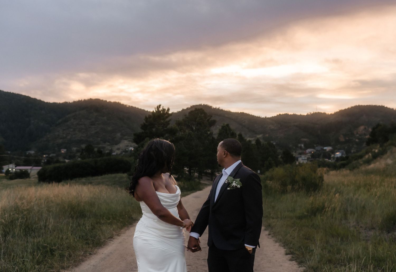Two people in tuxedos stand on beach with pampas grass and lantern wedding decor at sunset.