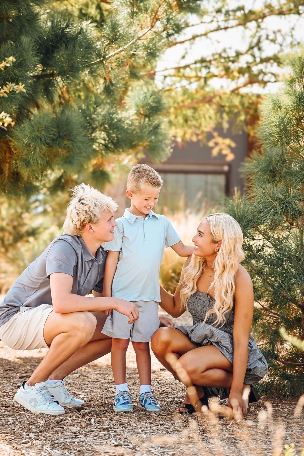A happy family with two blonde parents crouching beside their young son outdoors among pine trees in warm sunlight.