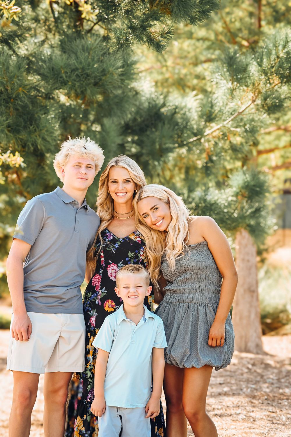 Family of four posing outdoors near pine trees, featuring a mother in floral dress with two teens and a young boy.