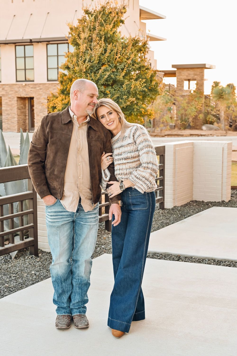 A couple smiles warmly while standing together outdoors near a modern brick building surrounded by autumn foliage.