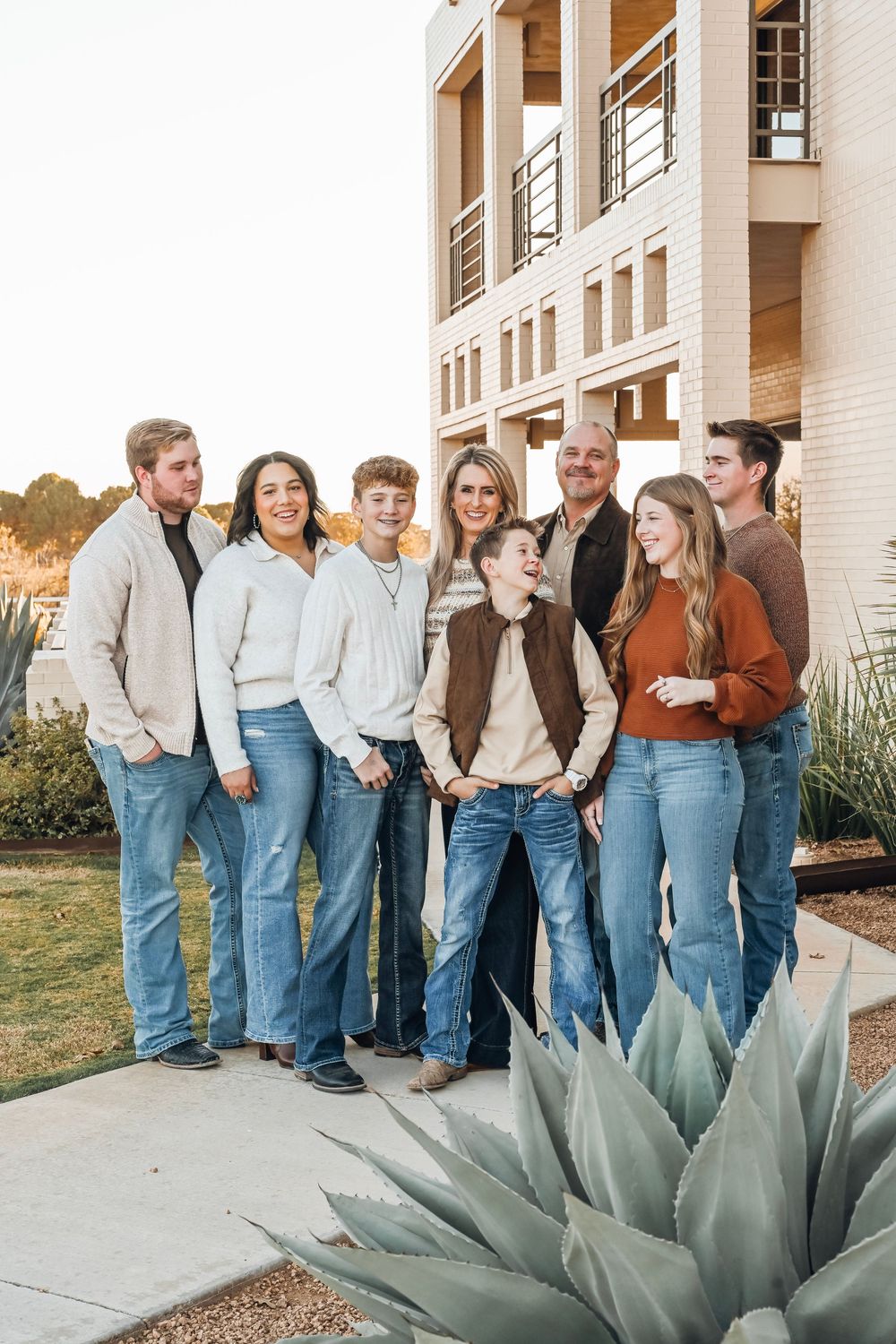 Large family group portrait outdoors in jeans and casual tops, posing near modern building with agave plant in foreground.