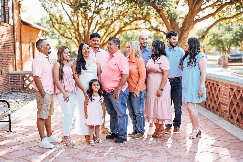 Large family group portrait outdoors on brick patio, wearing coordinated pastel outfits, smiling together in warm golden sunlight.
