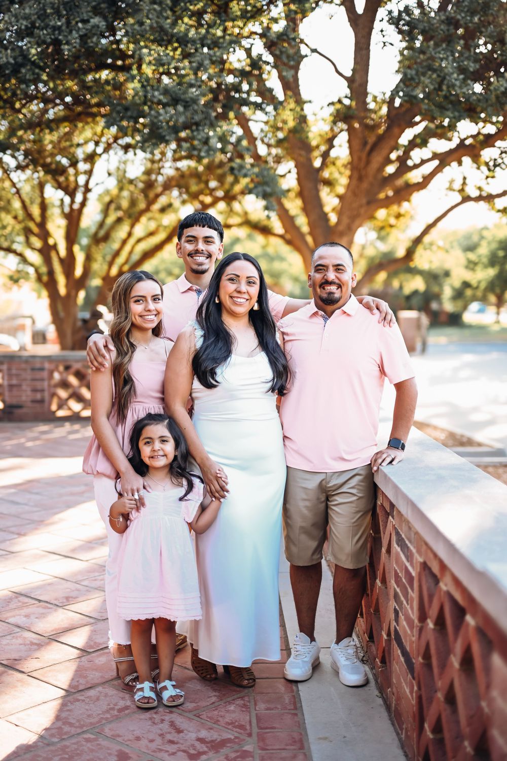 Family of five posing outdoors under large trees, wearing coordinated pink and white outfits on a sunny day.