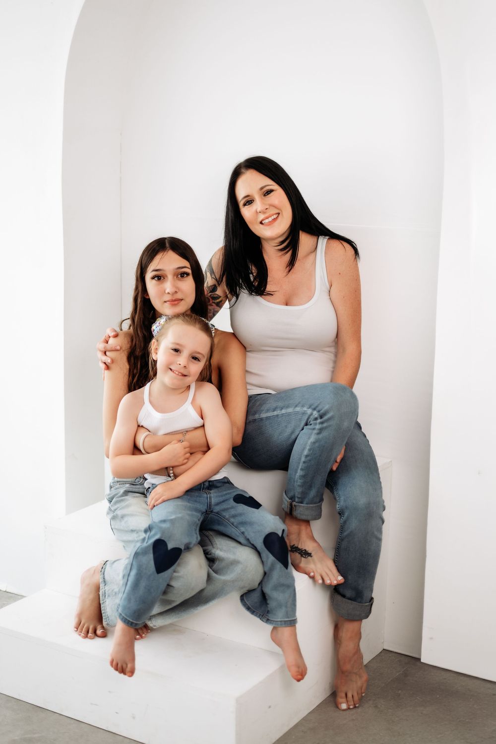 A woman and two girls in jeans and white tops pose together on white steps in a bright studio setting.