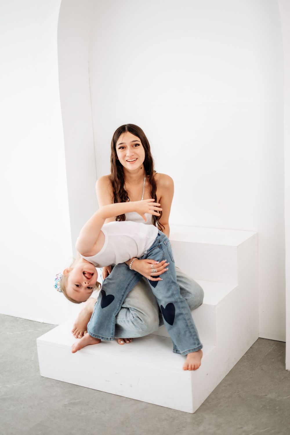 A smiling teenage girl sits on white steps holding a laughing toddler upside down in a bright white room.