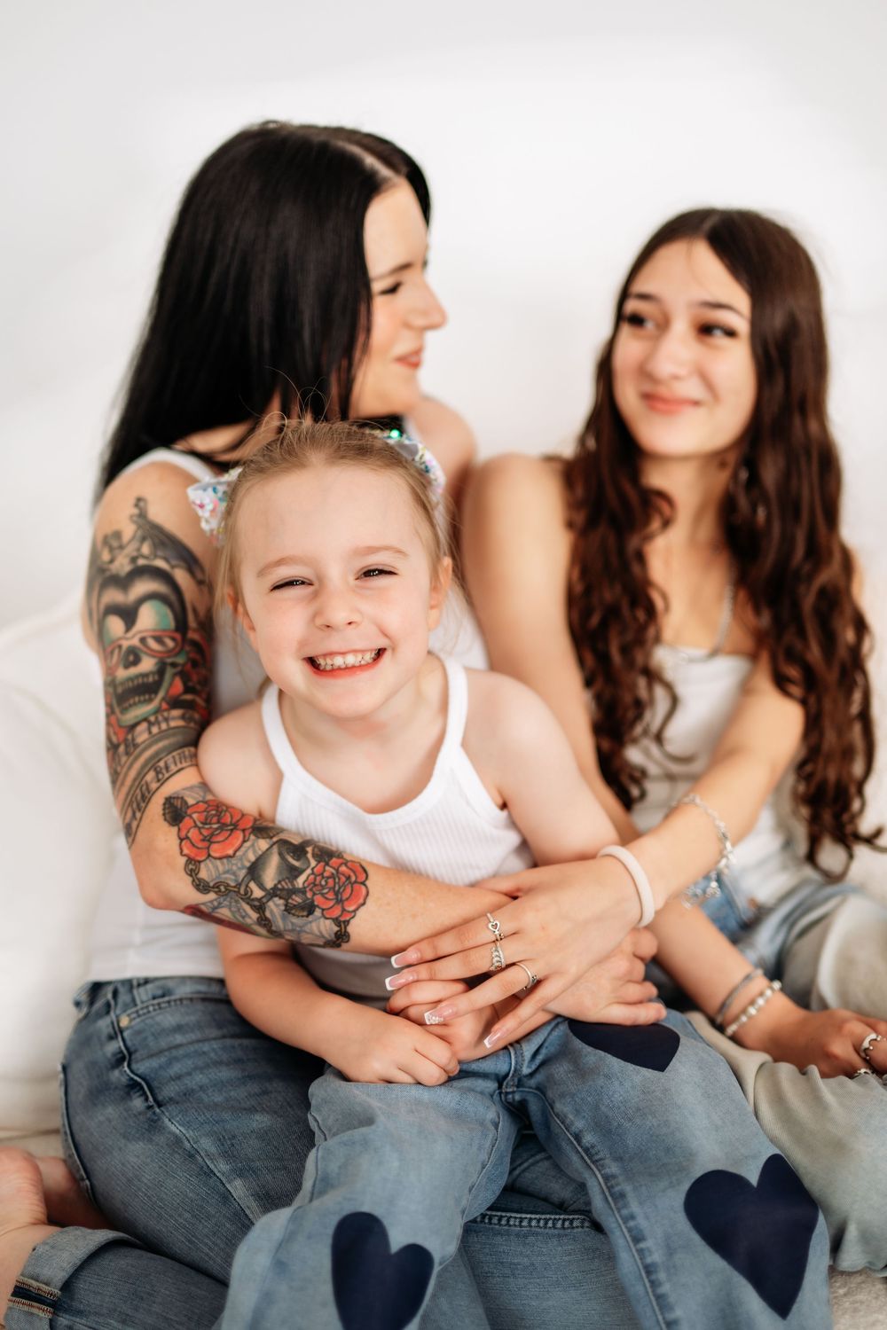 A tattooed woman smiles while hugging a young blonde girl, with a teenage girl sitting beside them on a white background.