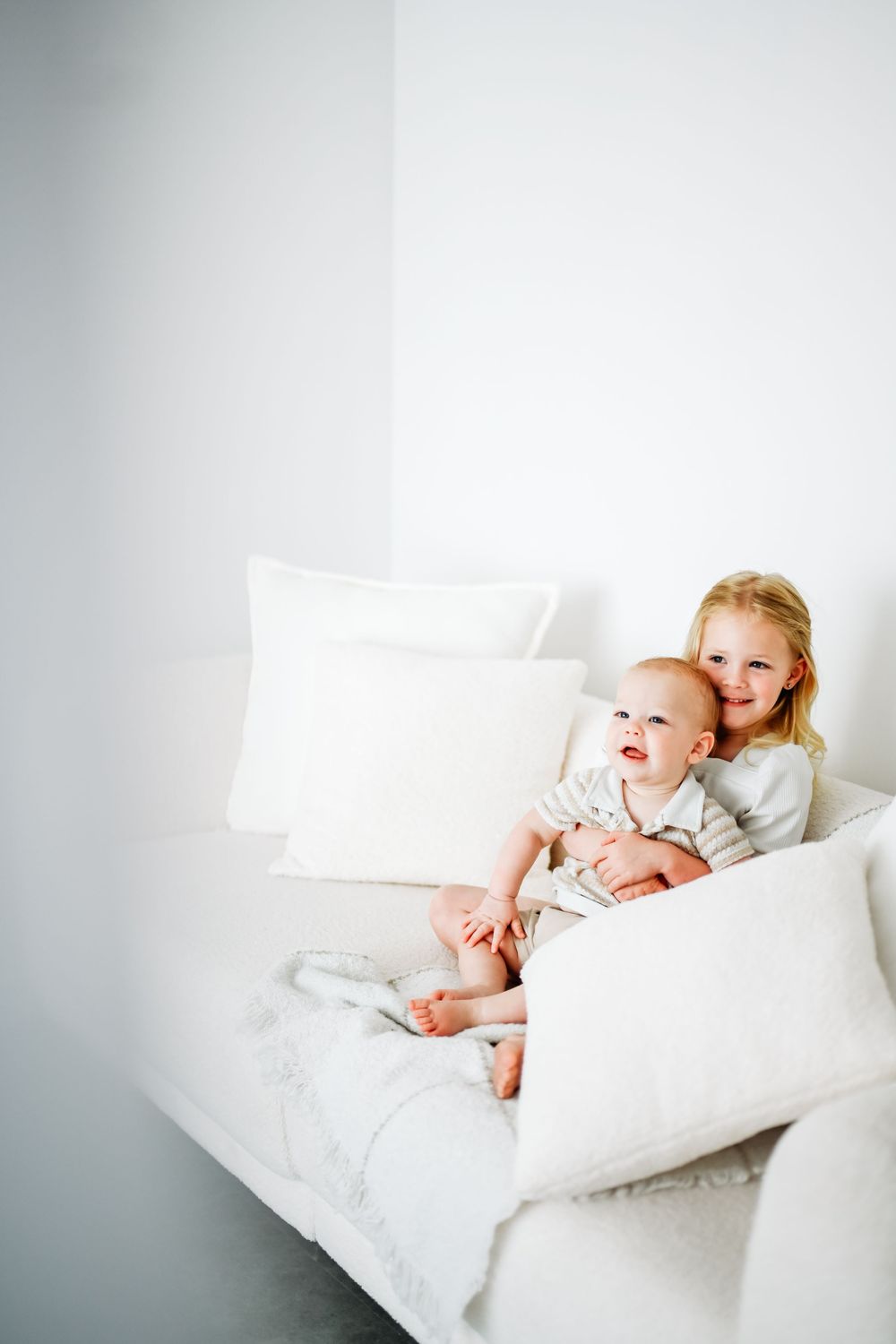 Young girl smiling and hugging baby sibling on white couch with pillows in bright minimalist room.