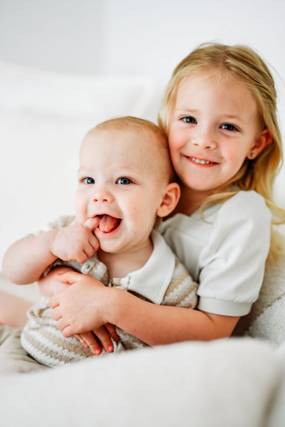 A smiling blonde girl hugs a happy baby sibling, both dressed in light colors on a white background.