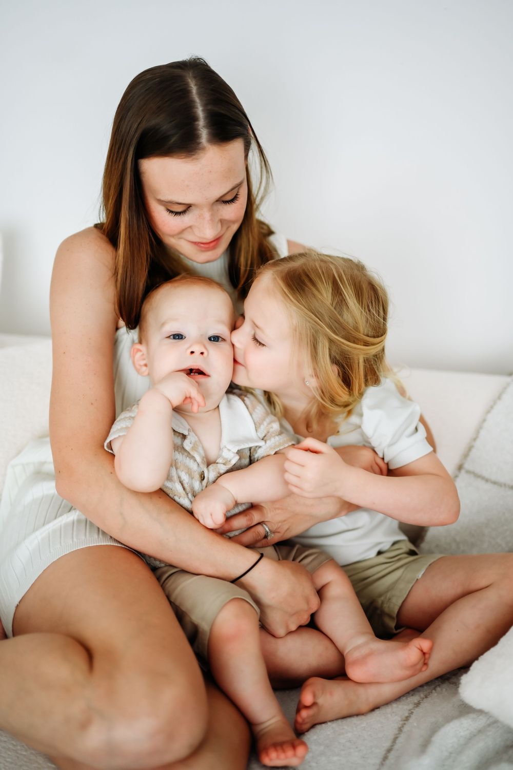 A young mother sits on a bed smiling as her toddler kisses a baby sibling in a warm family moment.