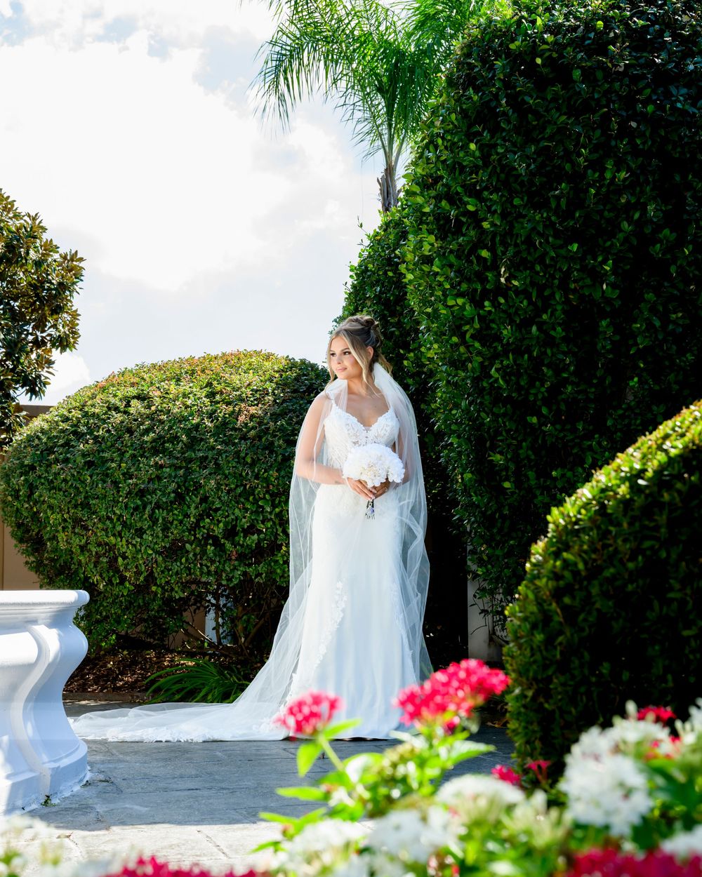 A person in a white lace wedding dress and veil stands elegantly in a garden setting with pink flowers and greenery.
