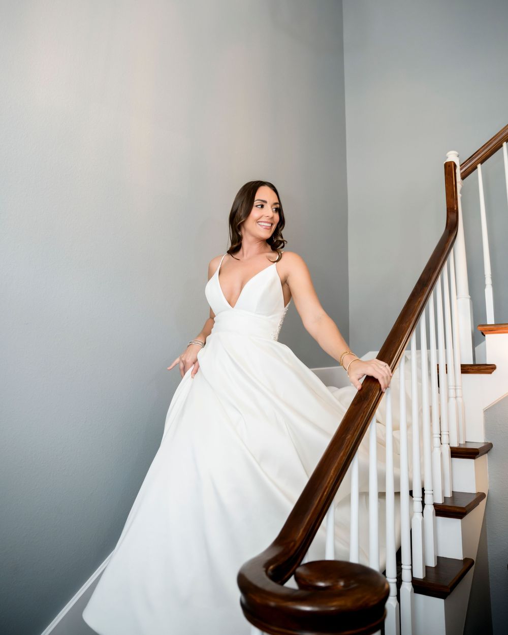 Bride in flowing white dress poses dramatically on staircase with wooden banister against grey wall.