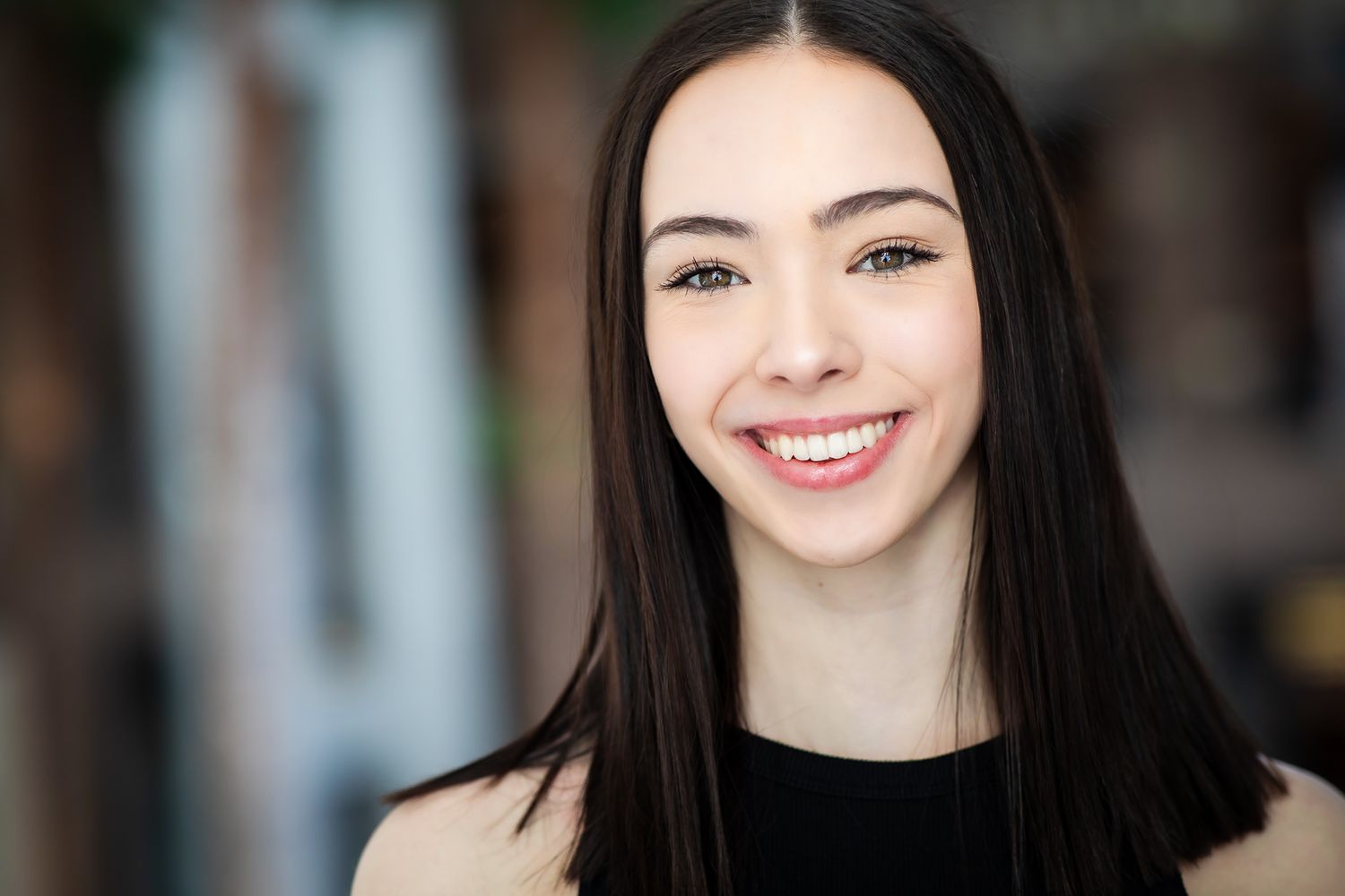 Young woman with long dark hair and bright smile wearing black sleeveless top in indoor setting.