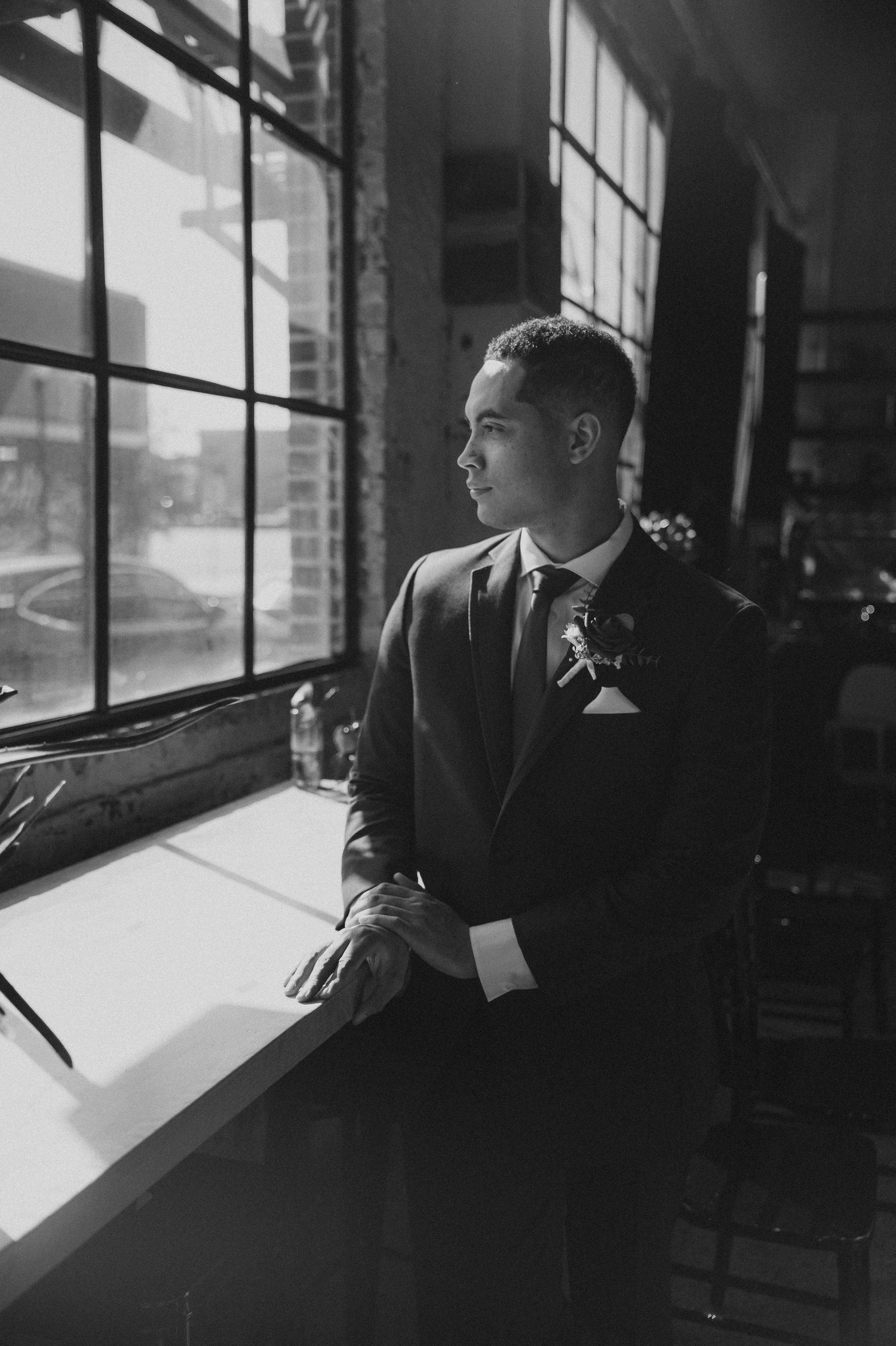 Moody black and white portrait of a groom gazing through industrial windows.
