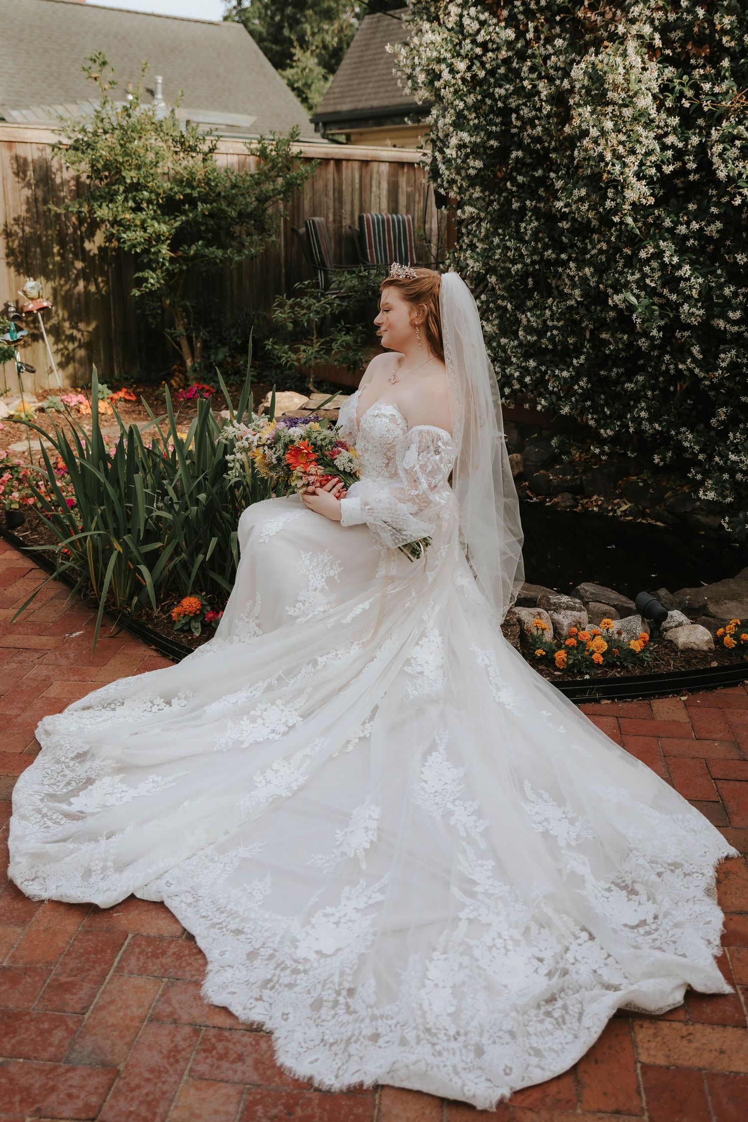 Stunning lace wedding dress with flowing train displayed in a garden setting with orange flowers.