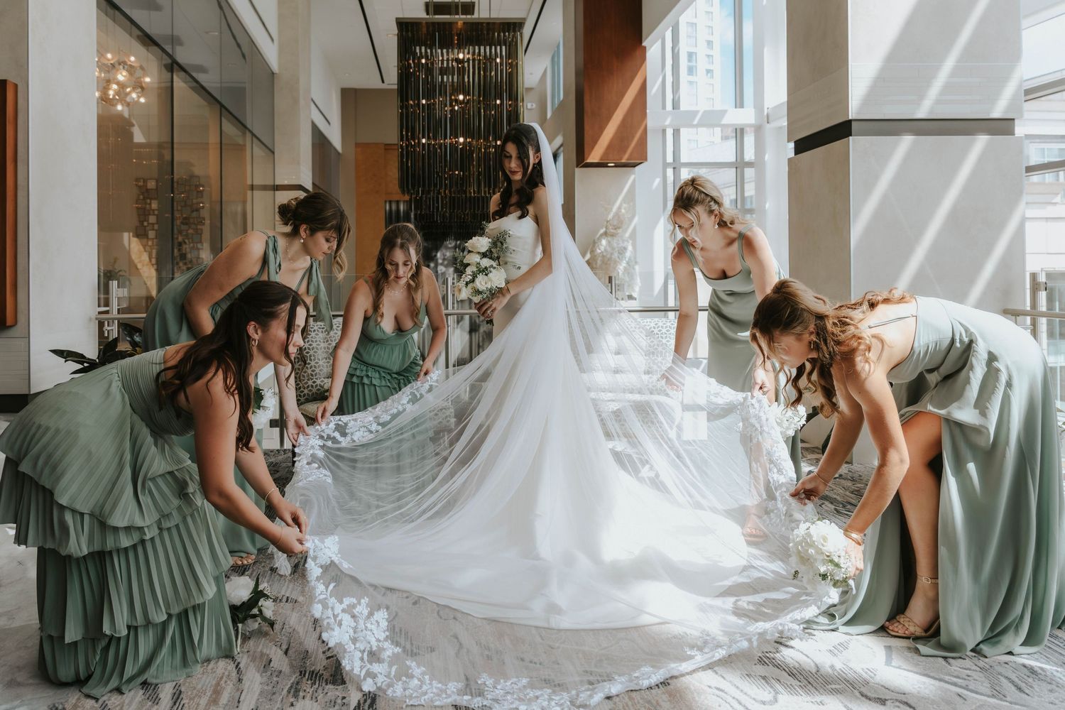 Bridesmaids in sage green dresses help arrange a flowing white wedding gown train in an elegant hallway.