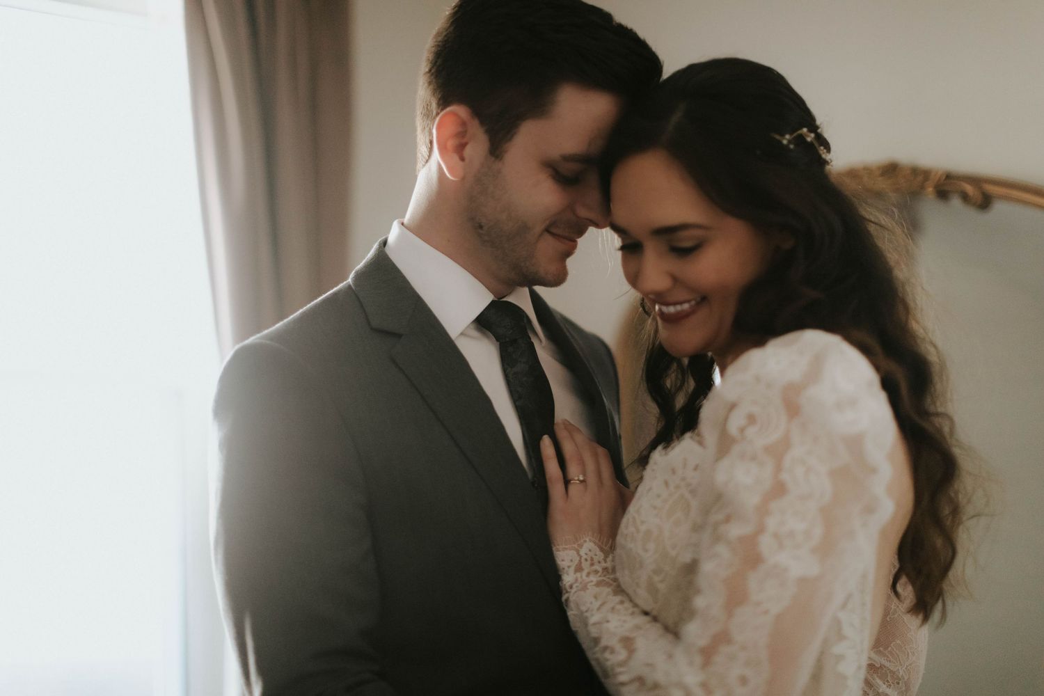 A bride and groom share an intimate forehead touch, smiling softly in warm backlit light on their wedding day.